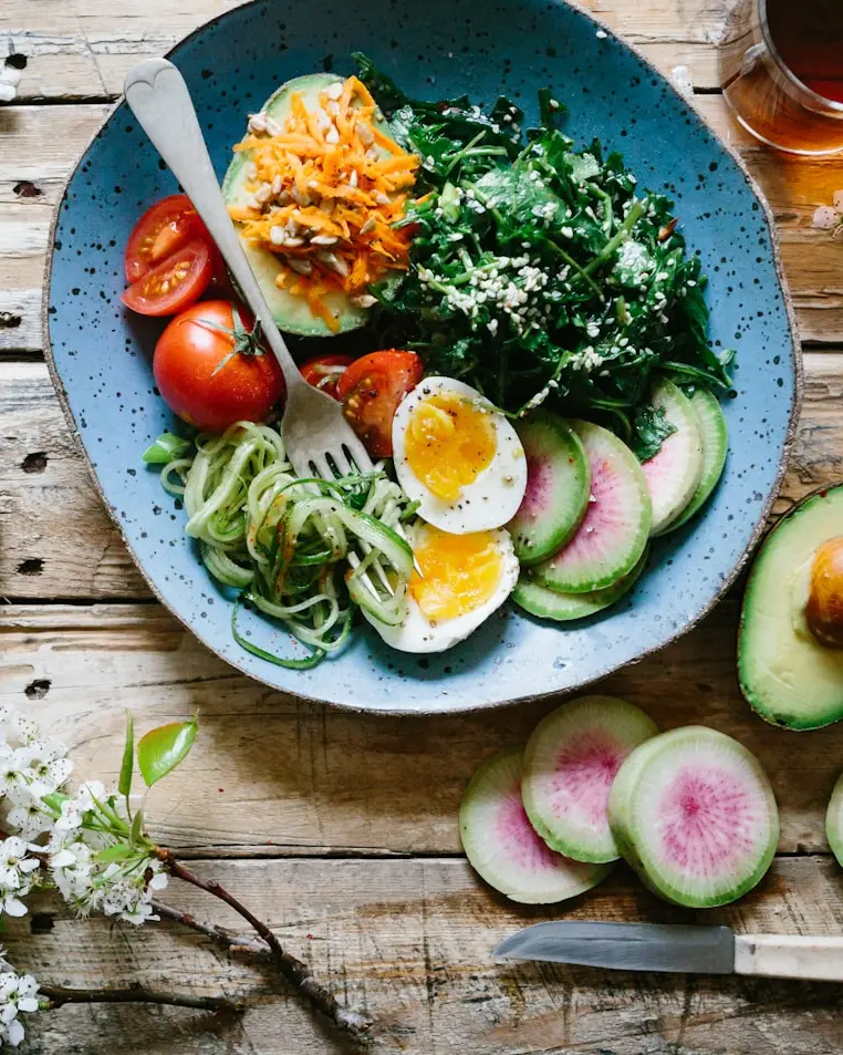 A bowl of greens, sliced tomato, courgette noodles and a soft-boiled egg on a wooden table, with an avocado and radishes beside it.